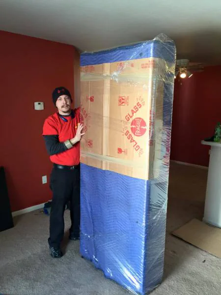 A mover in a red shirt and black knit cap stands indoors beside a tall piece padded with blue moving blankets and cartons marked GLASS and FRAGILE, all sealed in clear stretch wrap