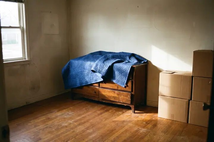 A wooden dresser just set down against the wall of an empty bedroom, its blue moving blanket half pulled off, taped cartons stacked beside it and late sun on the wall and the hardwood floor